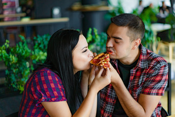 A pretty young couple eating one slice of pizza together. A nice girl and handsome man biting the same pizza in a modern cafe.