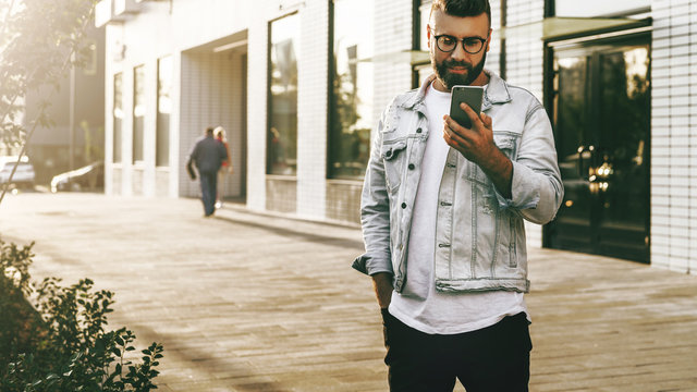 Handsome smiling hipster businessman with beard, in denim jacket and trendy glasses walks around city and using smartphone.Happy guy is standing outdoor and checking email on digital gadget.Lifestyle.