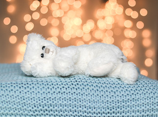 Fototapeta na wymiar Cute white little teddy bear lying peacefully on light blue knitted blanket. Cozy sweater weather. Soft warm pastel plaid with natural bokeh at background. Cold winter season.
