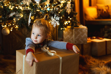 Fototapeta na wymiar A beautiful little girl hugs a box with a Christmas gift in front of a decorated Christmas tree. Family friendly Christmas concept.