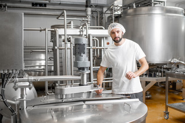 Portrait of a handsome worker in uniform near the stainless tank full with fermenting milk at the...