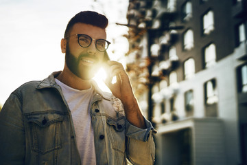 Handsome hipster businessman with beard, in denim jacket and trendy glasses walks around city and calls on mobile phone.