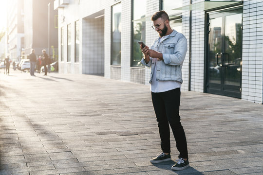 Handsome smiling hipster businessman with beard, in denim jacket, trendy glasses walks around city and using smartphone.