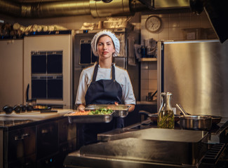 Female chef wearing a uniform holding a container with chopped vegetables at restaurant's kitchen.