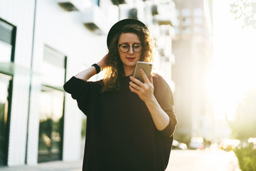 Young curly woman in hat and fashionable glasses makes photo on digital camera of smartphone, standing on city street.