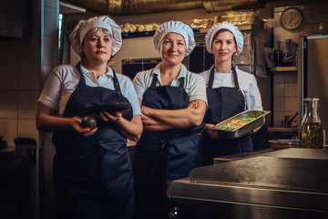 Cooking team wearing a uniform holding containers with chopped vegetables, posing for a camera at restaurant's kitchen.