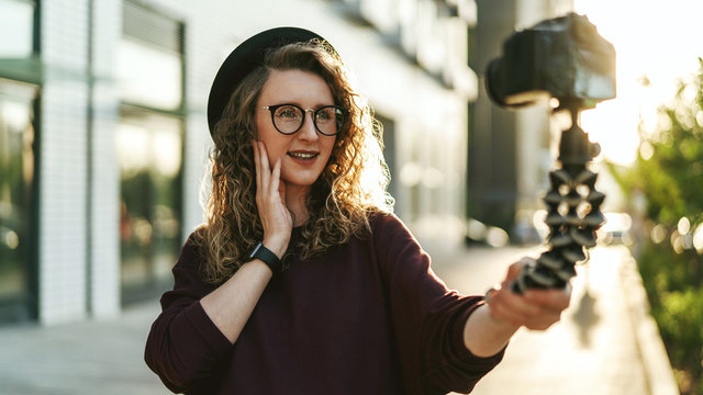 Young female blogger in trendy glasses and hat walks on city street, records herself on video holding camera for tripod.