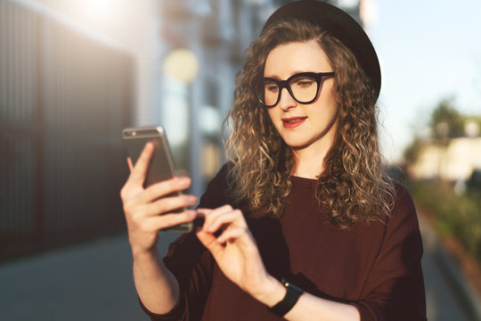 Young curly hipster woman in trendy glasses and hat walks on city street and uses smartphone. Social media,lifestyle.