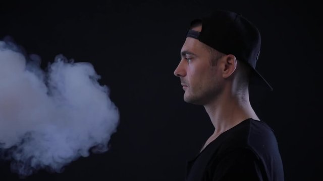 Smoking habit - the concept of exposure to others. Man smokes in the face. Man smoking electronic cigarette, vape in dark clothes. vapor on black isolated background.