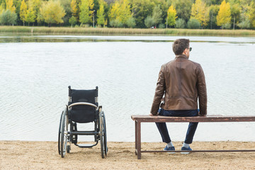 A young man sits on a bench by the lake, next to his wheelchair.