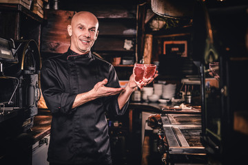 Smiling chef presenting a fresh steak before cooking and looking at a camera in a restaurant kitchen.