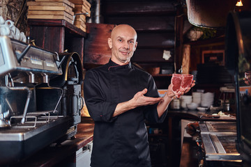 Confident chef presenting a fresh steak before cooking and looking at a camera in a restaurant kitchen.
