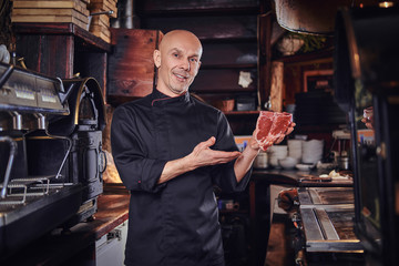 Naklejka premium Smiling chef presenting a fresh steak before cooking and looking at a camera in a restaurant kitchen.