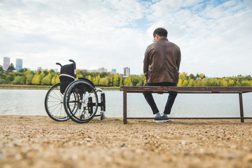 A young man sits on a bench by the lake, next to his wheelchair.