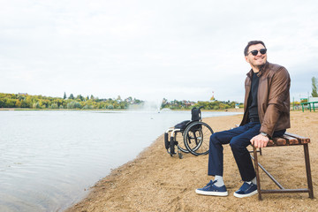 Obraz premium A young man sits on a bench by the lake, next to his wheelchair.