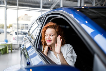 Red-haired girl sitting in the passenger seat of the car waving her hand from the window, outside...