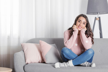Young woman in warm sweater sitting on sofa at home. Space for text