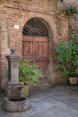 Fototapeta na wymiar Courtyard in the medieval Village of Buonconvento, Italy with a water fountain and plants surrounding a door