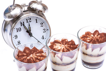 Diet concept with alarm clock: sweet Italian dessert tiramasu with delicate mascarpone cream from traditional recipe in glass isolated on white background