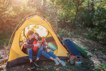 Lovely couple sitting in tent. She leans to him and keep eyes closed. Her shoulders are covered with blanket. Bearded guy embrace young woman. They hold cups.