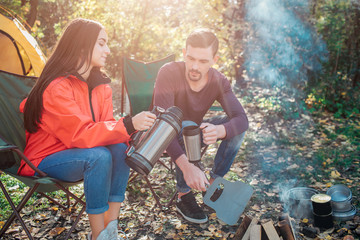Young woman sit on folding chair and poures hot water from thermos into man's cup. She does it accurate. Guy hold thermocup in hand. They sit at fire together.