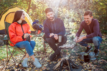 Friends sitting at fire and spending time together. Young man on right holds grill with sausages on fire. They are cooking and smoking. Another guy looks at it. Young woman smile.