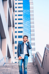 Young professional, wearing leather jacket, jeans, arm carrying laptop computer, talking to his family on his mobile phone, walking down stairs outside office building after working, going home.