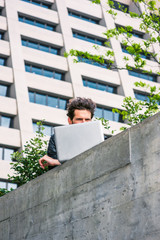 European graduate student studying in New York. A young guy, narrowing eyes, reading, working on laptop computer on the top of wall in business district in spring day. Power of Modern Reading.