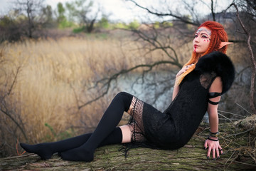 Elf woman with fiery hair on a log. Beautiful young fantasy style girl. Cosplay character