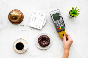 Pay the bill by payment terminal. Woman's hand insert bank card in payment terminal near bill, service bell, coffee on white stone background top view