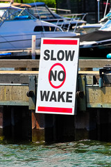 A slow no wake sign on a dock with boats in the background