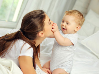 Mom and a charming blue-eyed baby of 10 months in a striped jumpsuit are lying in bed and hugging