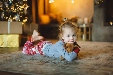 A beautiful baby lies on the floor in front of the tree and hugs her favorite plush toy