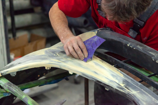 A Man Prepares A Car Body Element For Painting After An Accident With The Help Of Abrasive Paper In A Car Repair Shop. Recovery Bumper After A Collision.