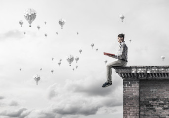 Man on roof edge reading book and aerostats flying in sky