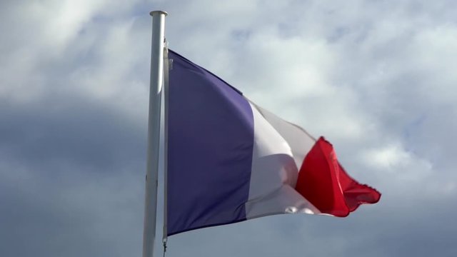 Slow motion of french flag waving in the wind on flagpole at an city. Slow-motion of France banner flutters at day in front of blue sky. Close up of a real fabric texture ensign fluttering-Dan