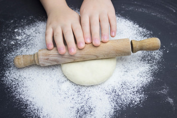 Kid's hands, some flour, wheat dough and rolling pin on the black table. Children hands making the rye dough for backing bread. Small hands kneading dough. Little child preparing dough for backing .