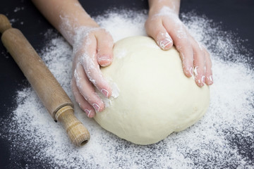 Kid's hands, some flour, wheat dough and rolling pin on the black table. Children hands making the rye dough for backing bread. Small hands kneading dough. Little child preparing dough for backing .