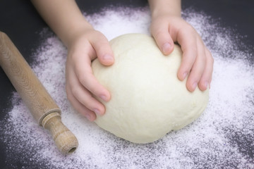 Kid's hands, some flour, wheat dough and rolling pin on the black table. Children hands making the rye dough for backing bread. Small hands kneading dough. Little child preparing dough for backing .
