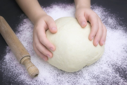 Kid's hands, some flour, wheat dough and rolling pin on the black table. Children hands making the rye dough for backing bread. Small hands kneading dough. Little child preparing dough for backing . - Powered by Adobe