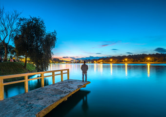 Fototapeta na wymiar Da Lat, Vietnam - October 29th, 2018: Man standing on a small bridge reflecting on the lake at sunrise as a relaxing way to welcome the beautiful new day