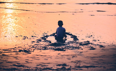 Silhouette of small  boy, kid, sitting alone on the beach at sunset time. kid sitting isolated on a beach view from behind, concept of solitude and alone.