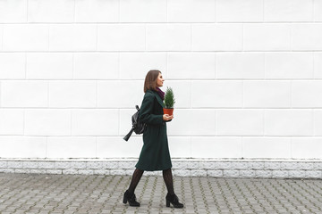 Beautiful young stylish woman wearing green coat, black high heel shoes and backpack walking through the city streets with small Christmas tree in her hands. New year concept. Christmas tree in a pot.
