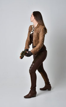 full length portrait of brunette  girl wearing brown leather steampunk outfit. standing pose, holding a gun. on grey studio background.