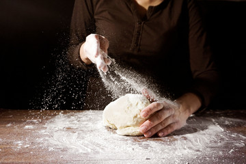 Cook hands kneading dough, sprinkling piece of dough with white wheat flour. Low key shot, close up on hands, some ingredients around on table.
