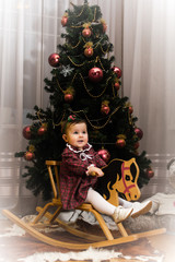 Little girl is sitting on wooden horse in front of Christmass tree