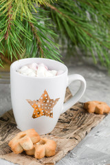 mug with drink decorated with marshmallow and star shape cookies  near evergreen christmas tree branches gray concrete background