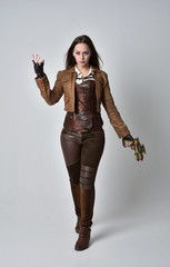 full length portrait of brunette  girl wearing brown leather steampunk outfit. standing pose, holding a gun. on grey studio background.
