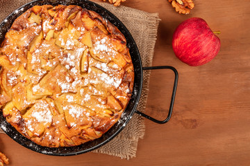 A closeup photo of an apple pie in a pan, shot from the top on a dark rustic wooden background with an apple, walnuts, and copy space