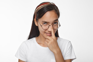 Studio image of playful mysterious young Asian woman with loose black hair adjusting her stylish round framed spectacles, looking at camera with suspicious or distrustful facial expression